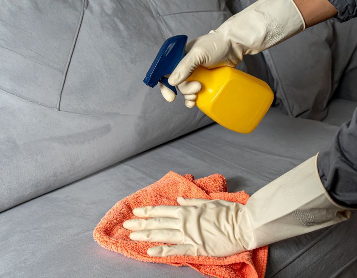 Person in gloves spraying and wiping a gray couch with a yellow spray bottle and orange cloth.