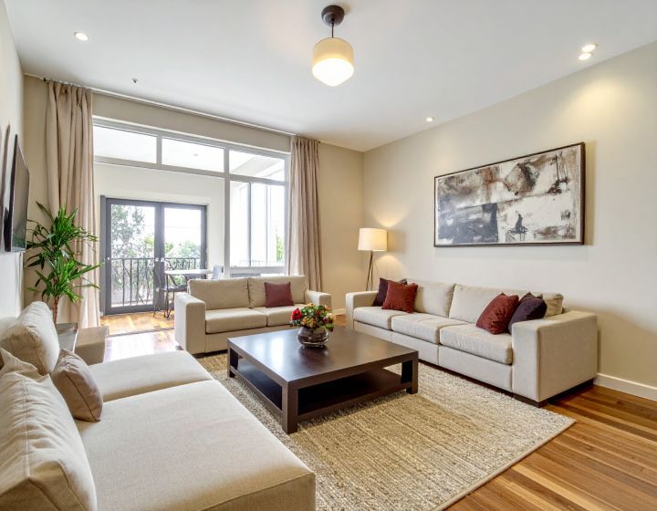 Living room with beige couches, dark wood coffee table, and large window overlooking a balcony.