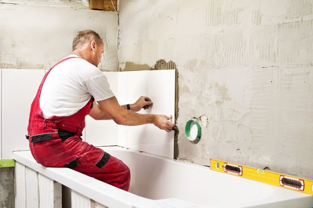 A Man Is Sitting On The Edge Of A Bathtub While Installing Tiles — Blueline Plumbing Services in Loganlea, QLD