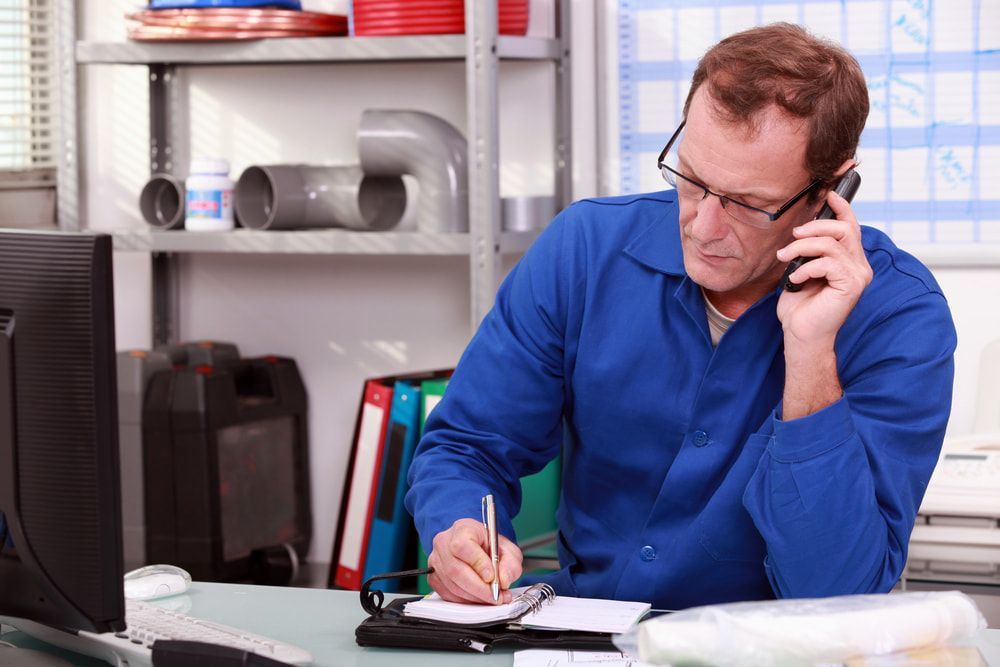 A Man In A Blue Shirt Is Sitting At A Desk Talking On A Cell Phone — Blueline Plumbing Services in Loganlea, QLD
