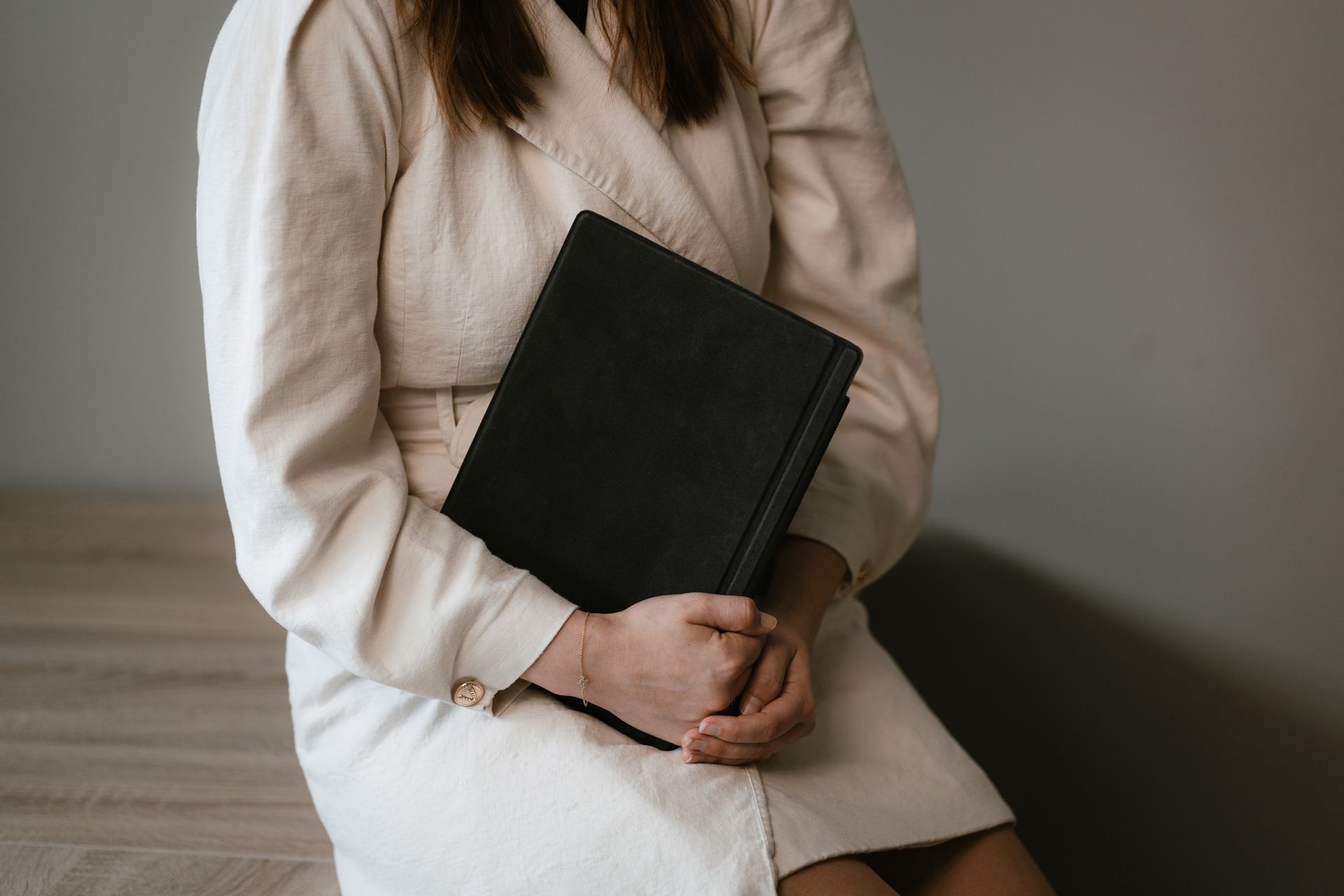 Woman in a beige coat sits, holding a closed black book.