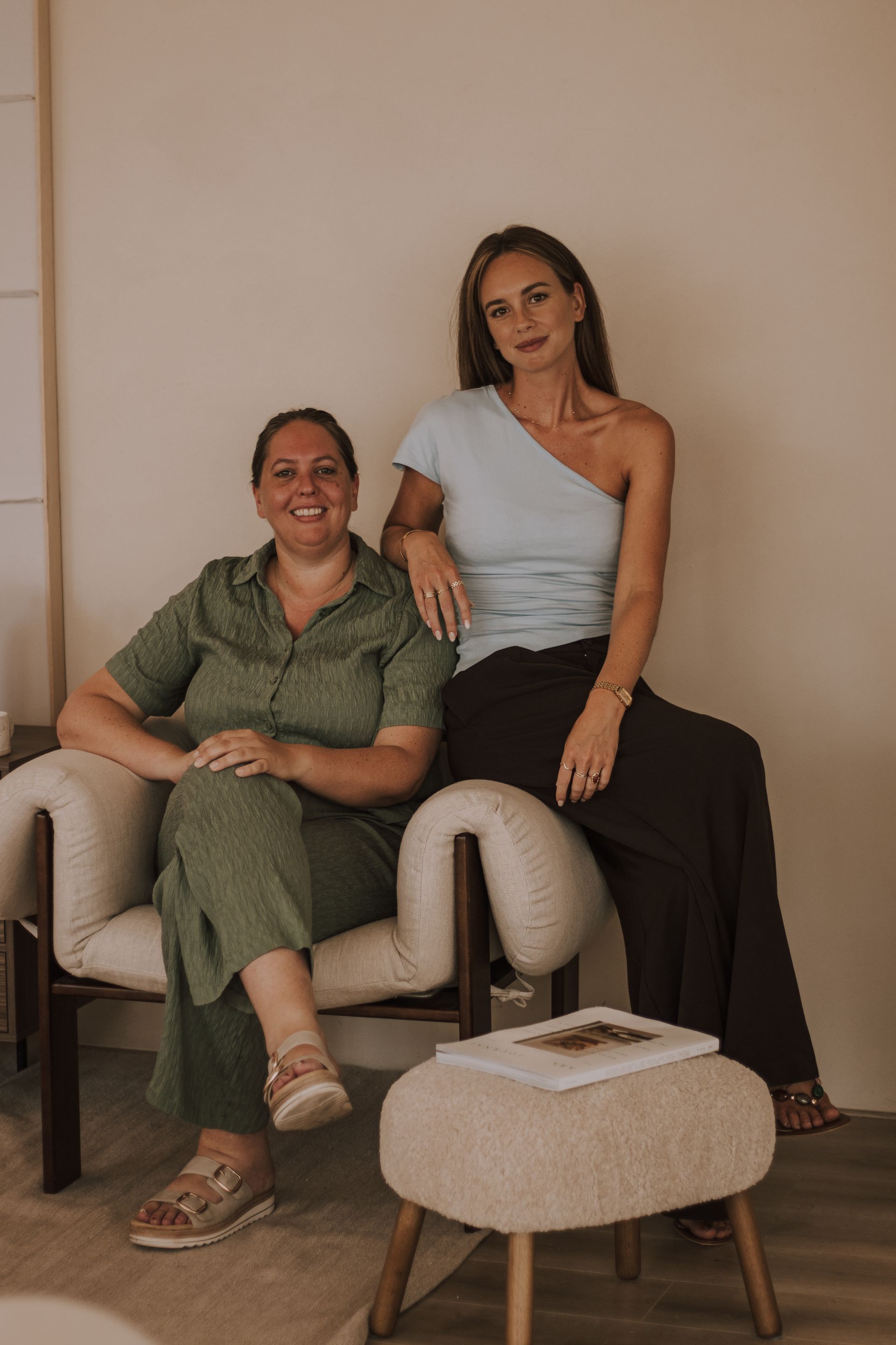Two women seated in a beige living room, one smiling in green, the other in a white top and black pants.