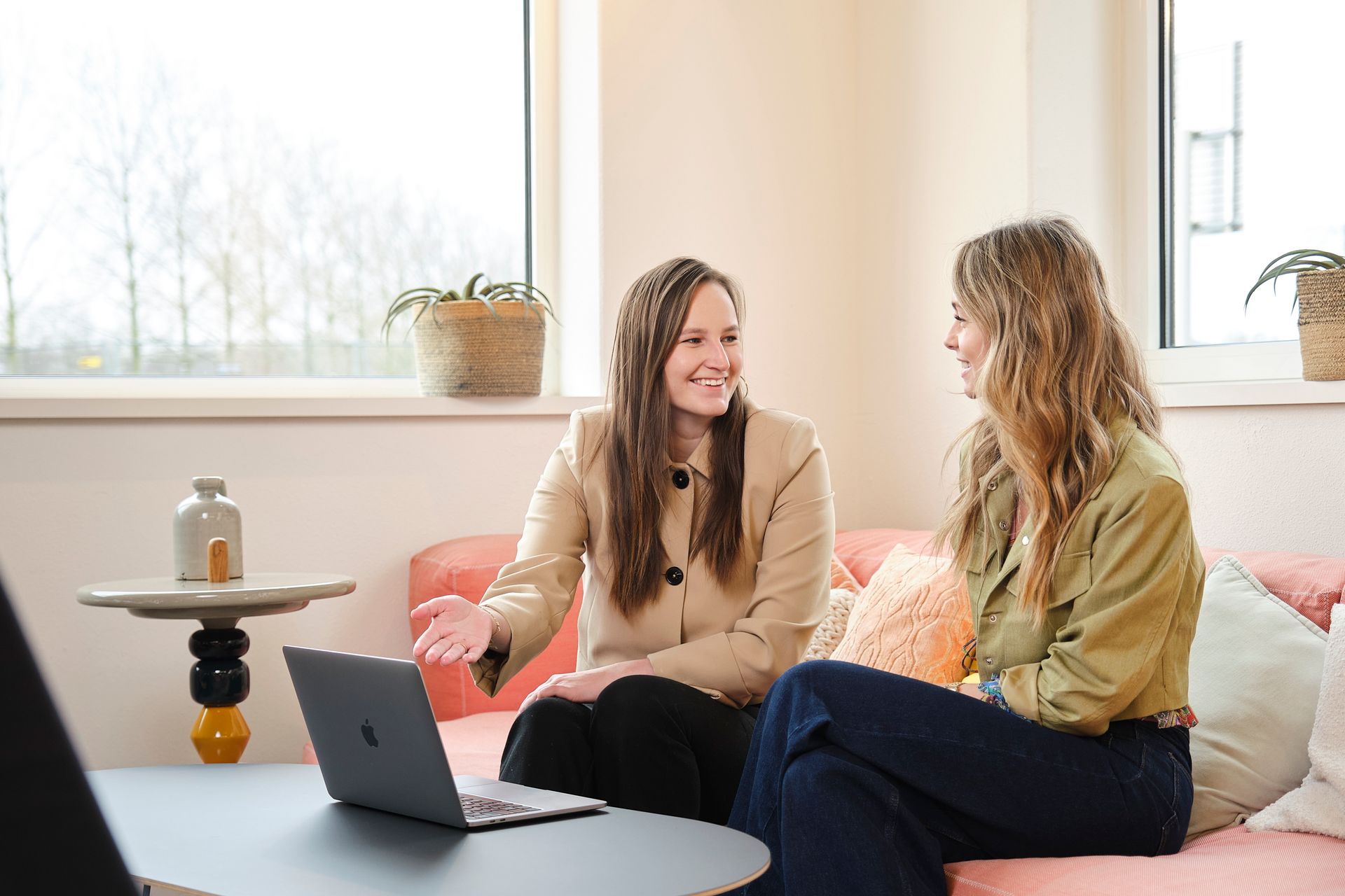 Two women talking on a pink couch beside a laptop in a bright living room