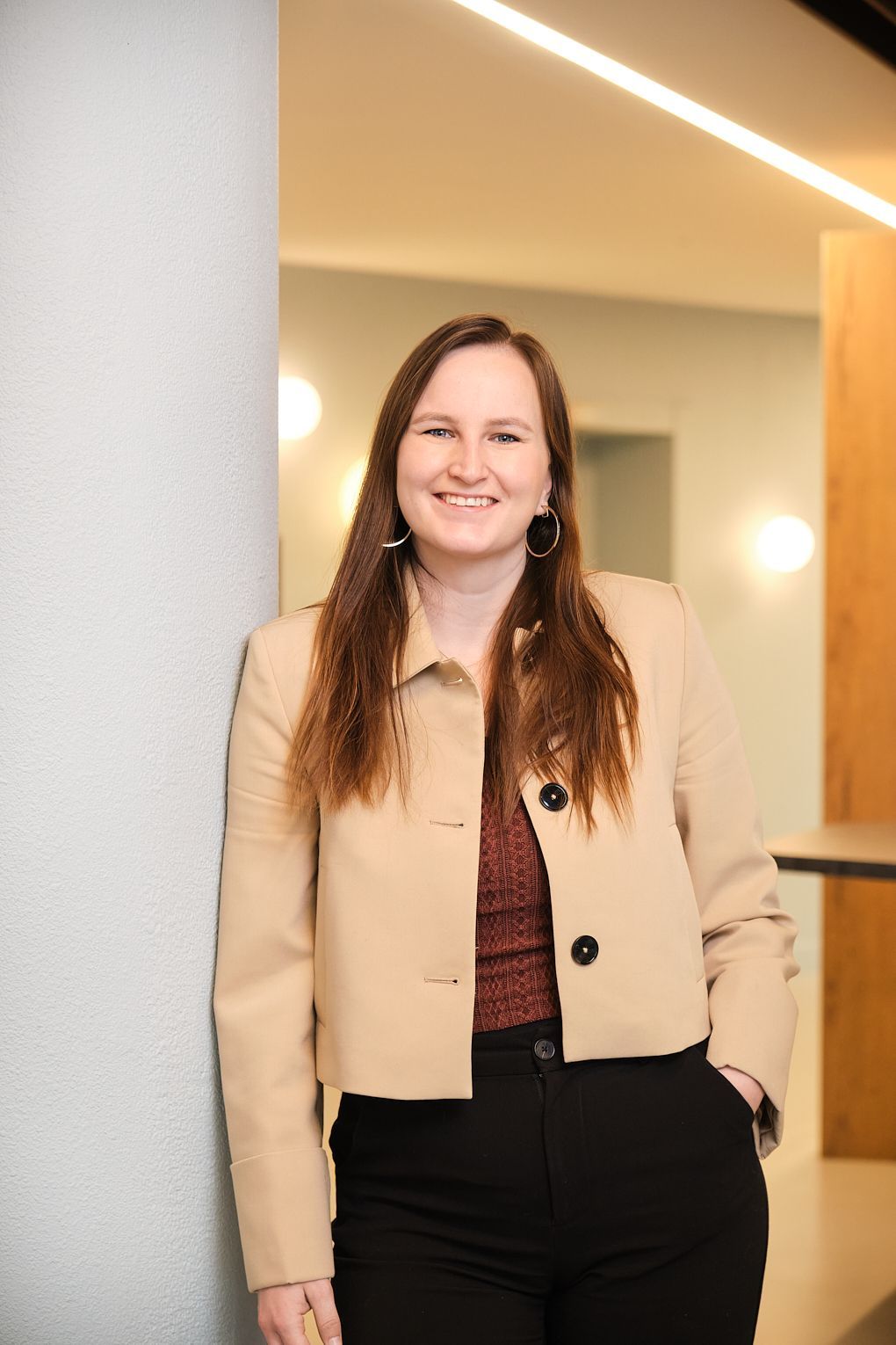 Een vrouw in een bruin jasje en een zwarte broek leunt tegen een muur.