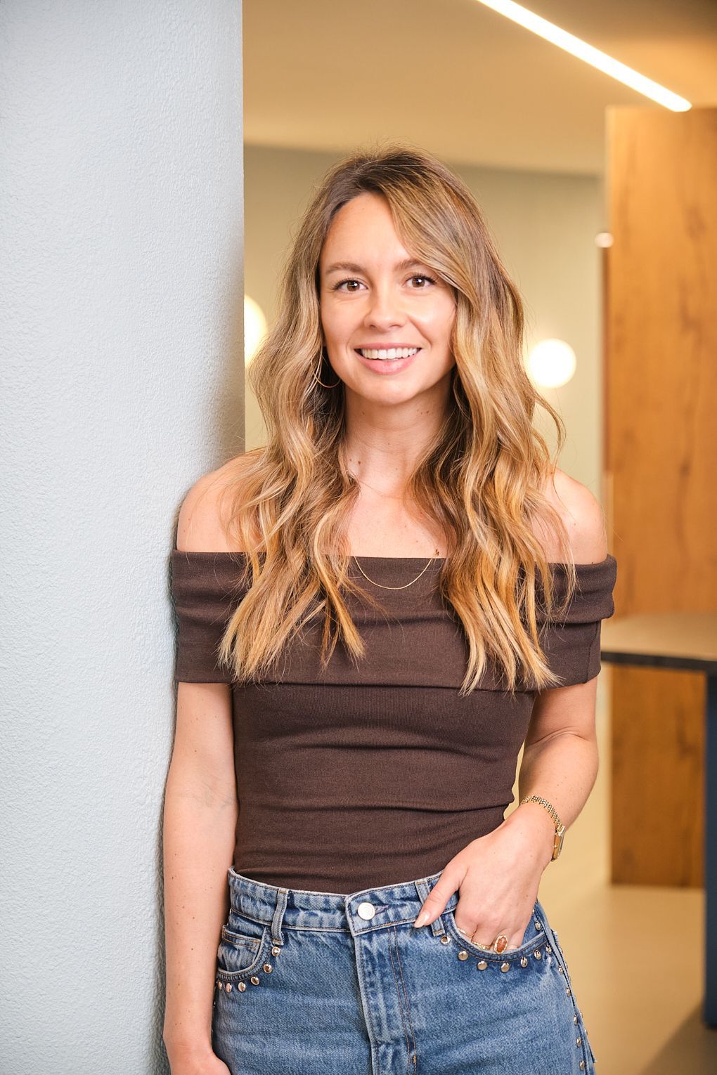 Een vrouw in een bruin topje met blote schouders en een blauwe spijkerbroek staat naast een muur.