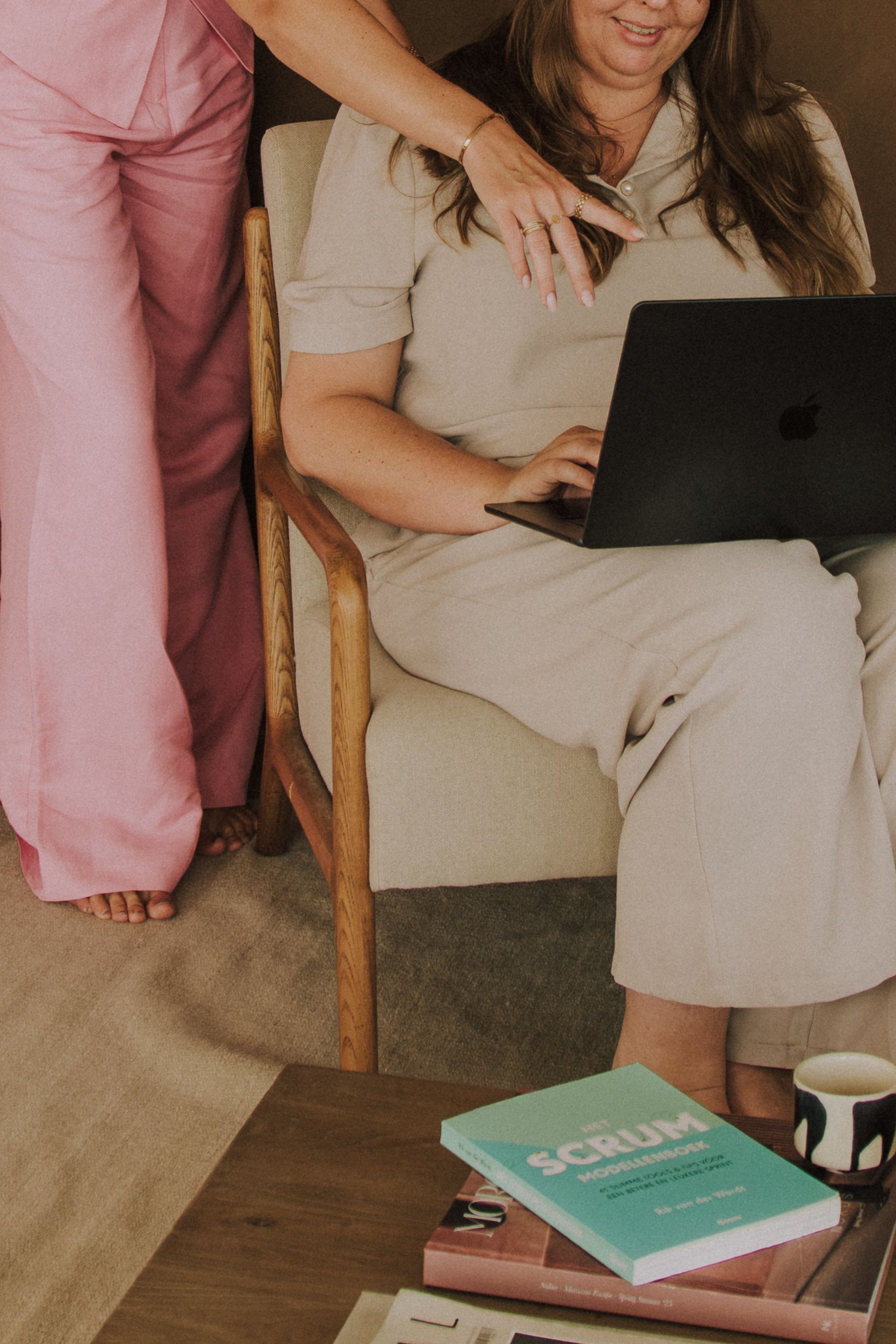Woman holding a laptop while seated on a chair; another person in pink stands beside her.