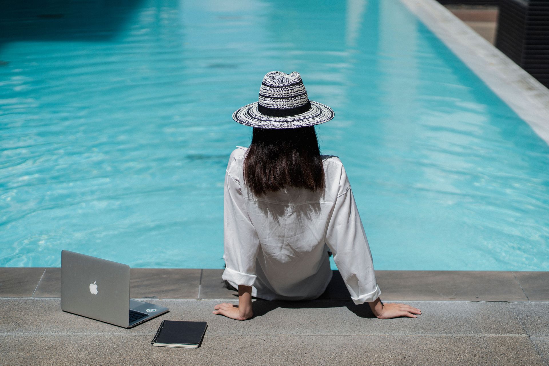 Een persoon met een zwart-witte hoed zit bij een turquoise zwembad naast een laptop.