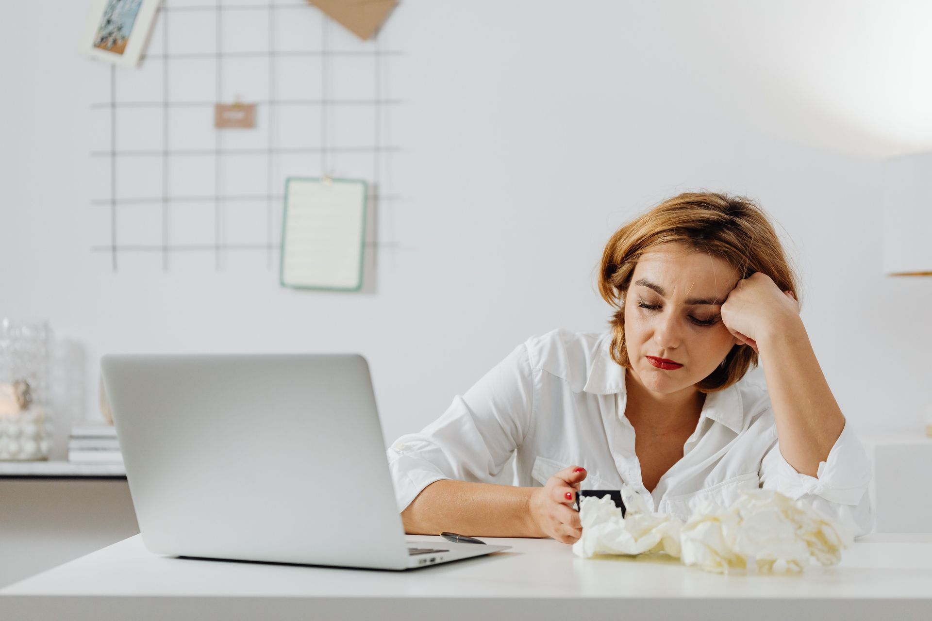 Woman at a desk with a laptop, resting her head on her hand, looking tired in a bright office.