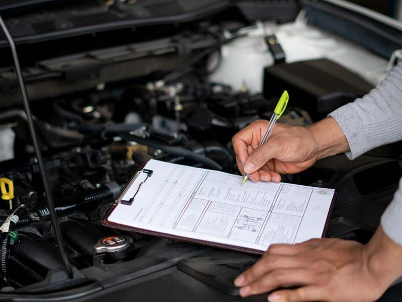 A person is writing on a clipboard in front of a car engine.