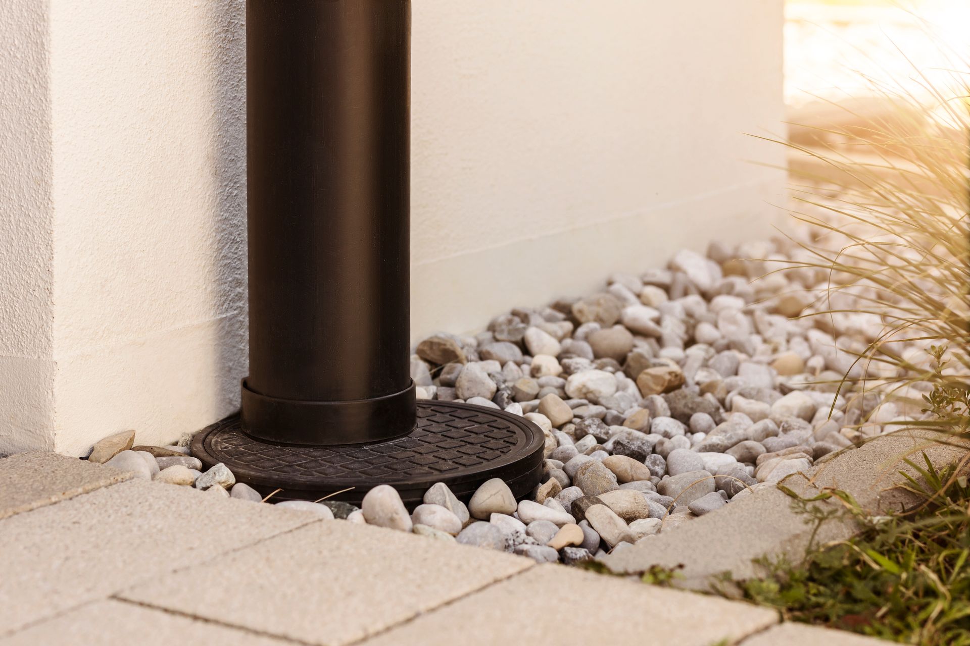 Black downspout on pebble bed beside a white wall and paved walkway