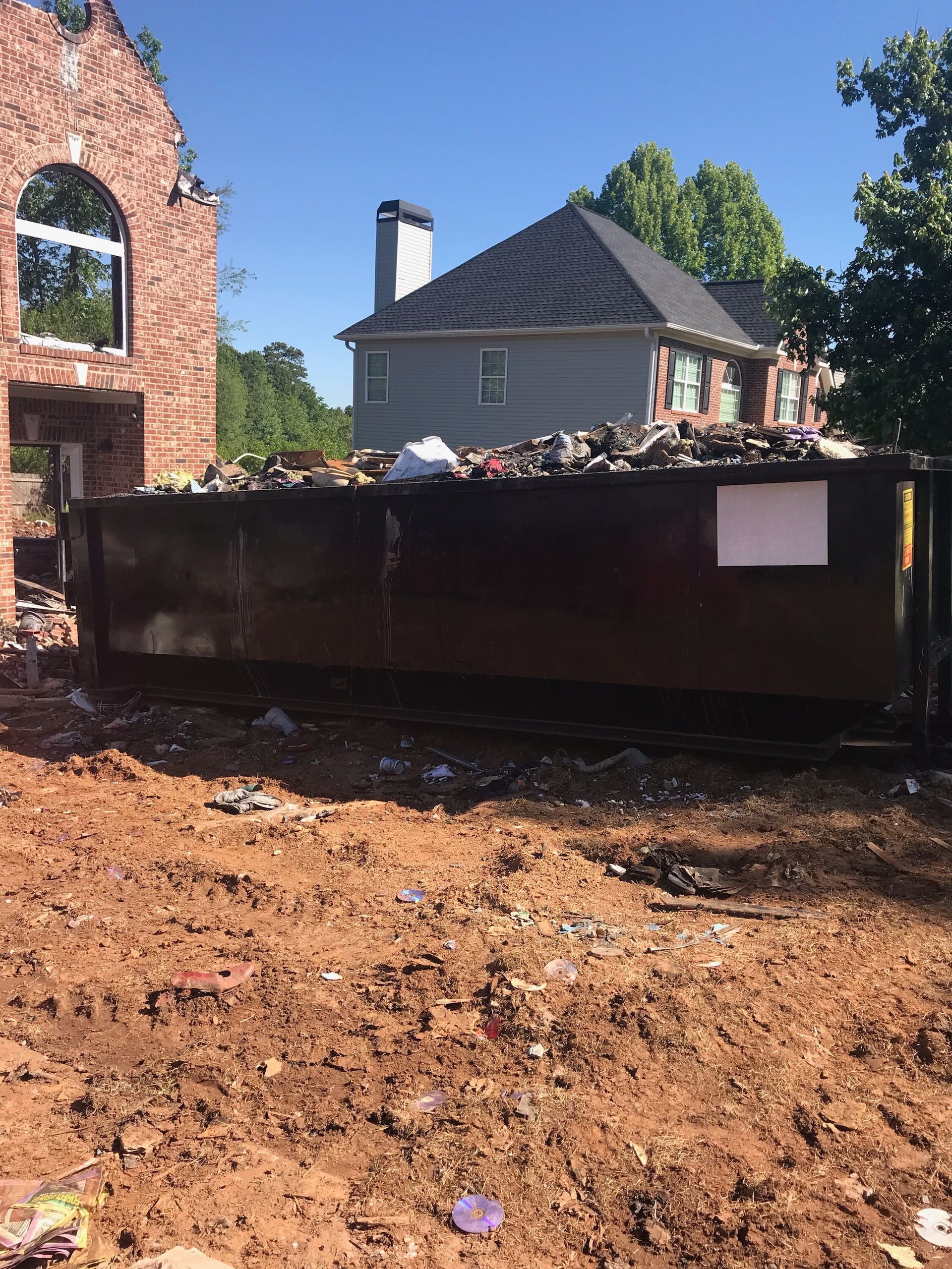 A dumpster is sitting in the dirt in front of a brick house.