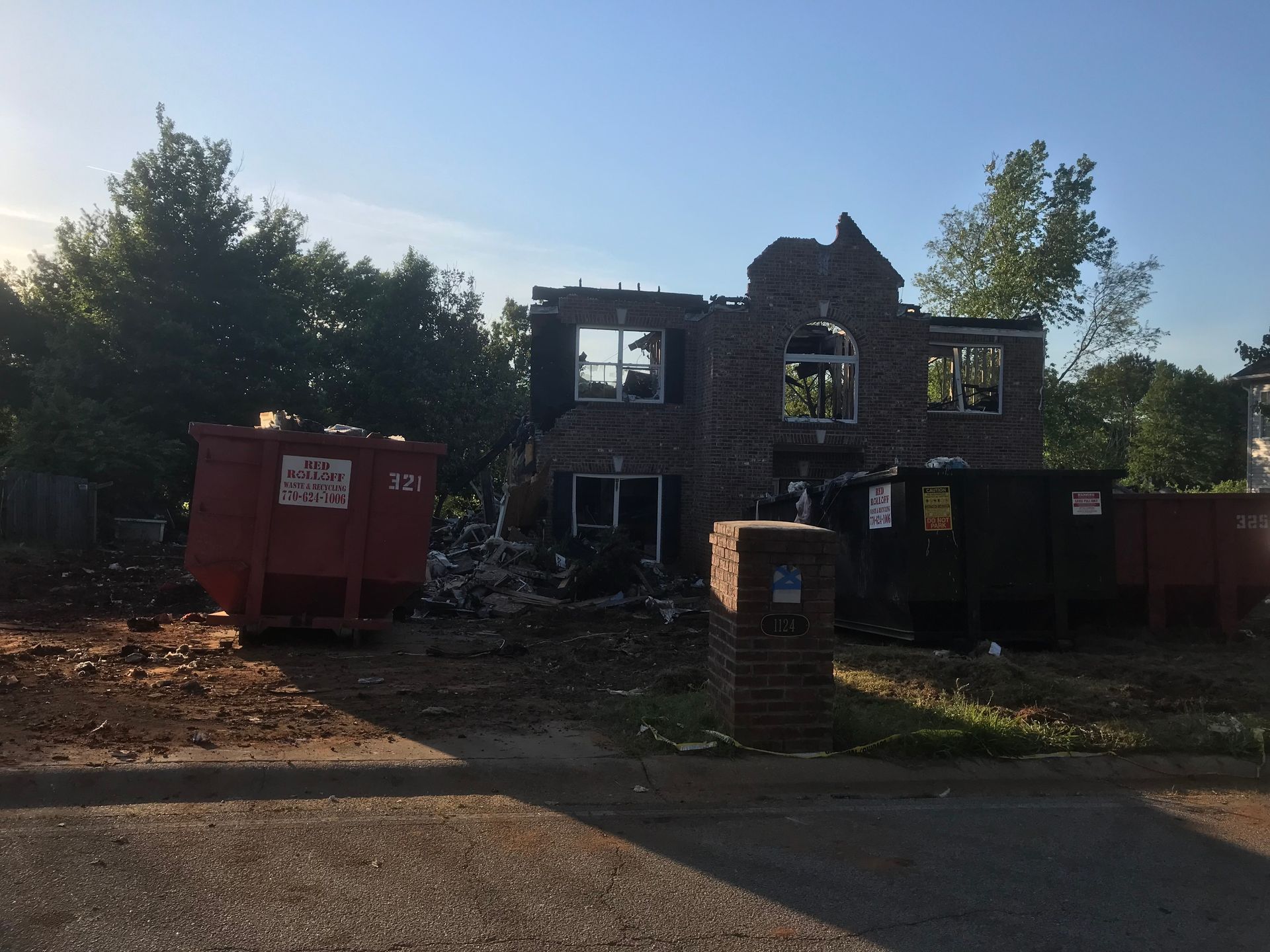 House destroyed by fire, with charred brick exterior, debris, and dumpsters.