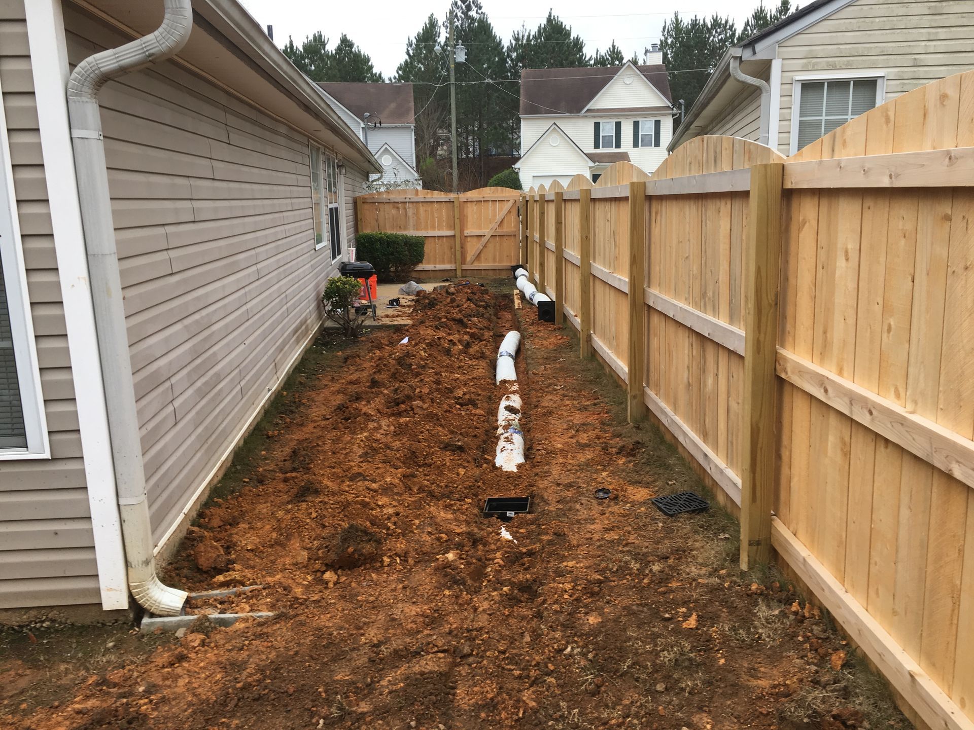A narrow dirt yard between a beige house and wooden fence, with a drainage pipe laid.