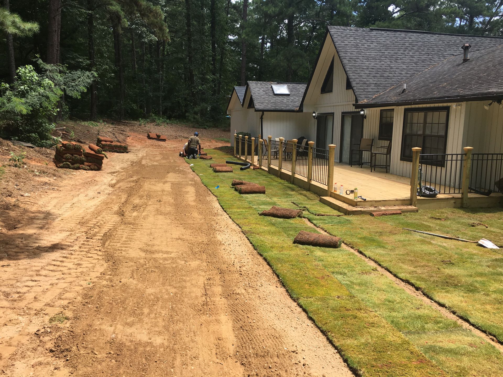 A person laying sod near a house with a deck, dirt path, and trees in the background.