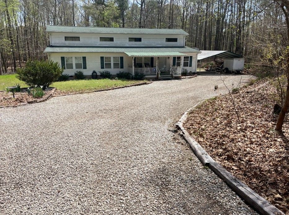 Two-story house with green roof and white siding, gravel driveway, and surrounding trees.