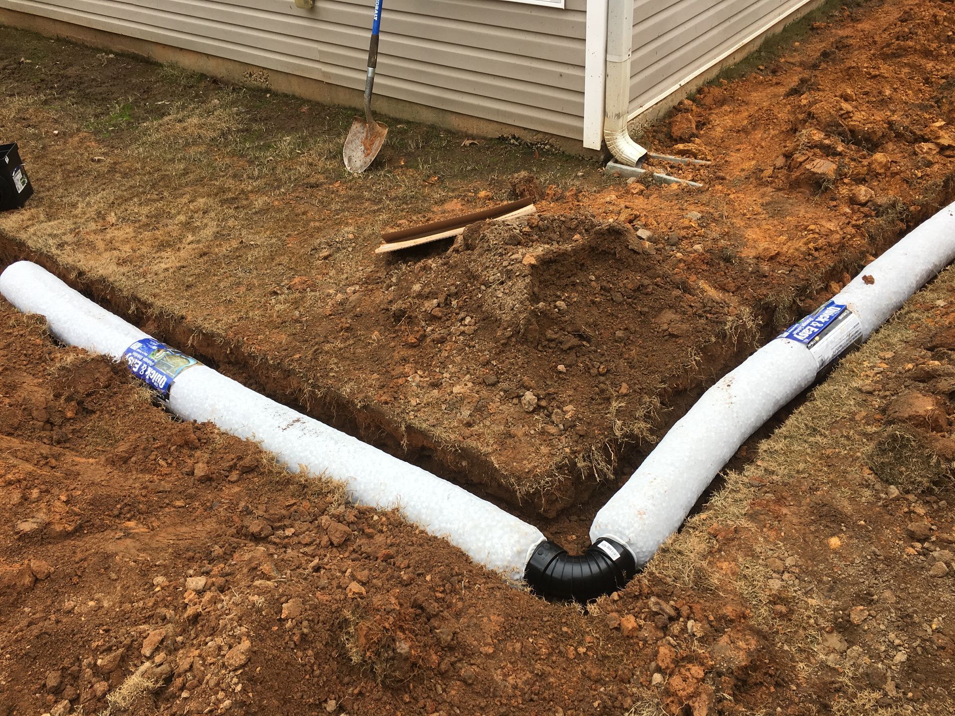 Trench with white drain pipe, black elbow, and soil being installed along the side of a building.