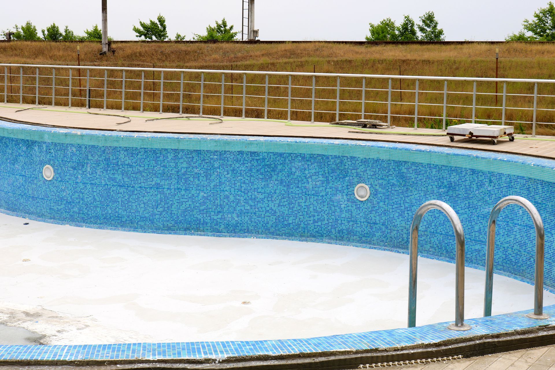 Empty blue-tiled swimming pool with ladder and metal railing, set against a grassy hill and overcast sky.