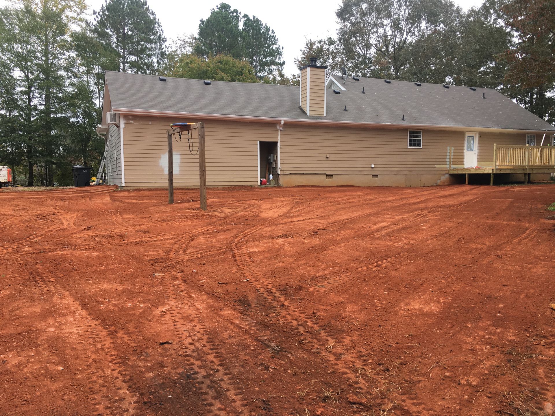 Rear view of a tan house with brown roof, surrounded by red dirt and trees.