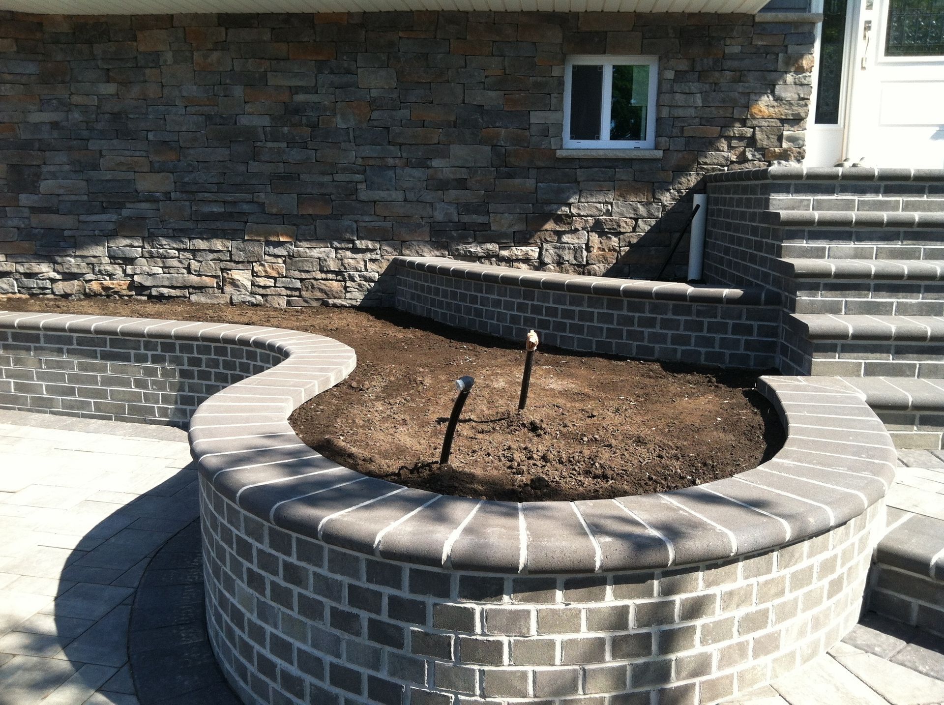 Curved brick planter filled with soil in front of a stone-clad house.