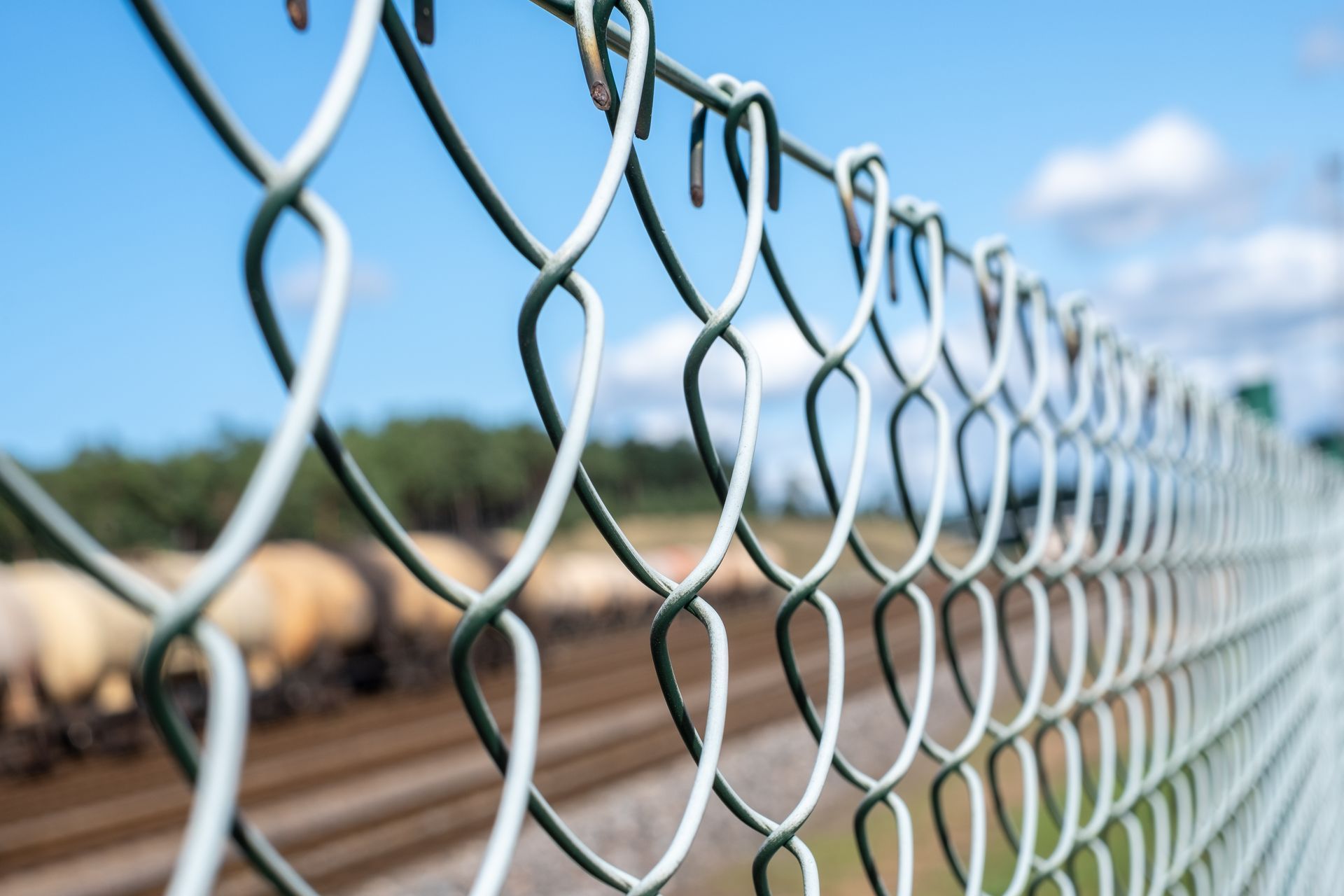 Chain-link fence in focus; blurred train cars and tracks in the background, blue sky.