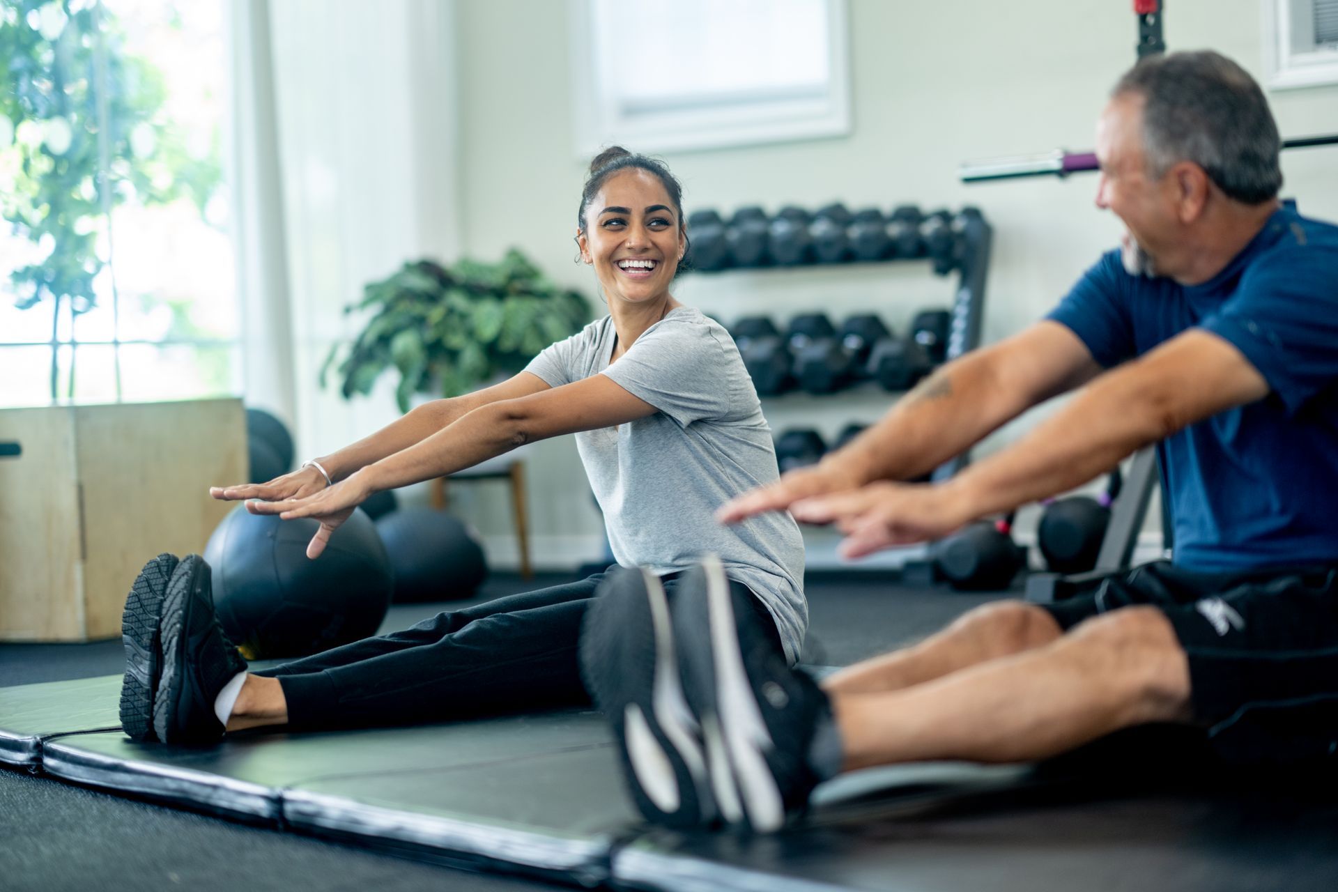 Una donna e un uomo sgranchiscono le gambe sul pavimento in palestra. Entrambi sorridono.