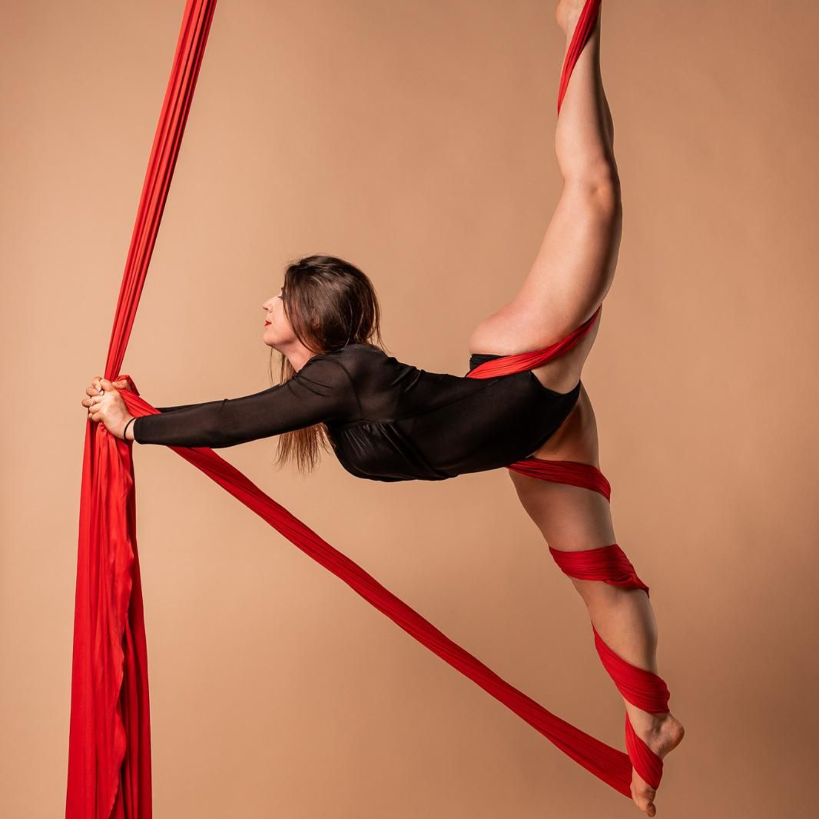 L'acrobata si inarca all'indietro, sospesa su teli rossi. Abiti scuri, riflettori puntati, fondale minimalista.
