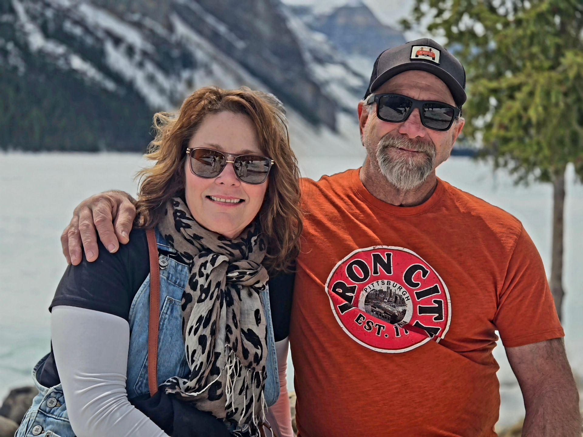 Two people smiling outdoors by a lake, with snowy mountains in the background.