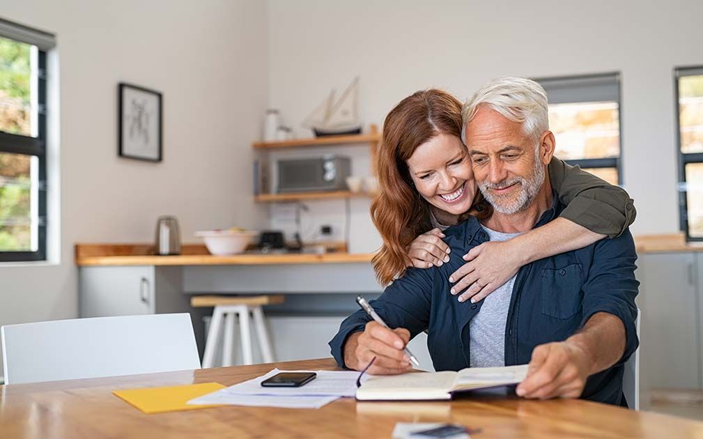 A woman is hugging a man while he writes in a notebook.