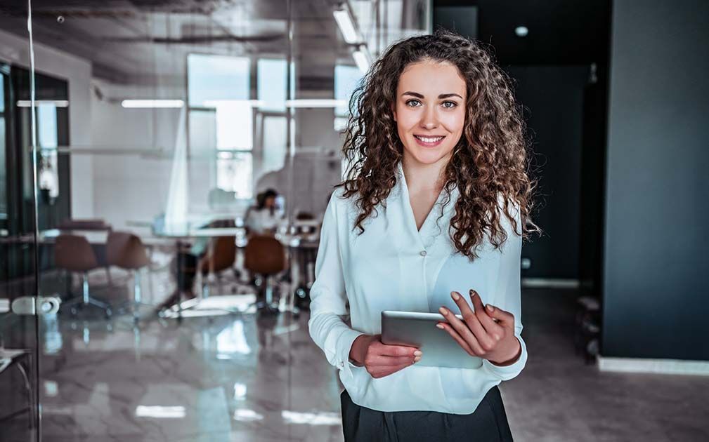 A woman is standing in an office holding a tablet computer.