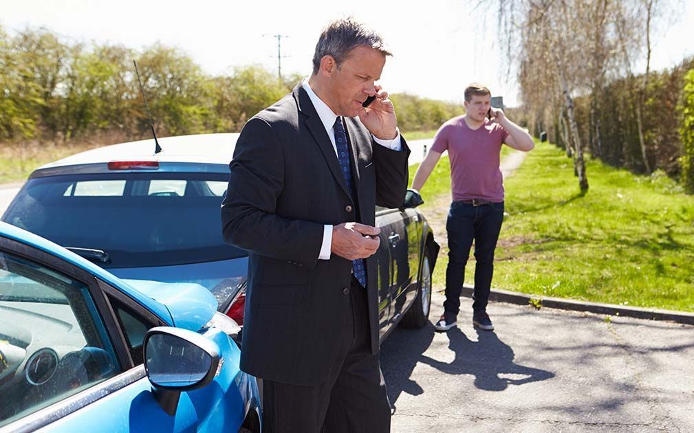 A man in a suit is talking on a cell phone next to a blue car.