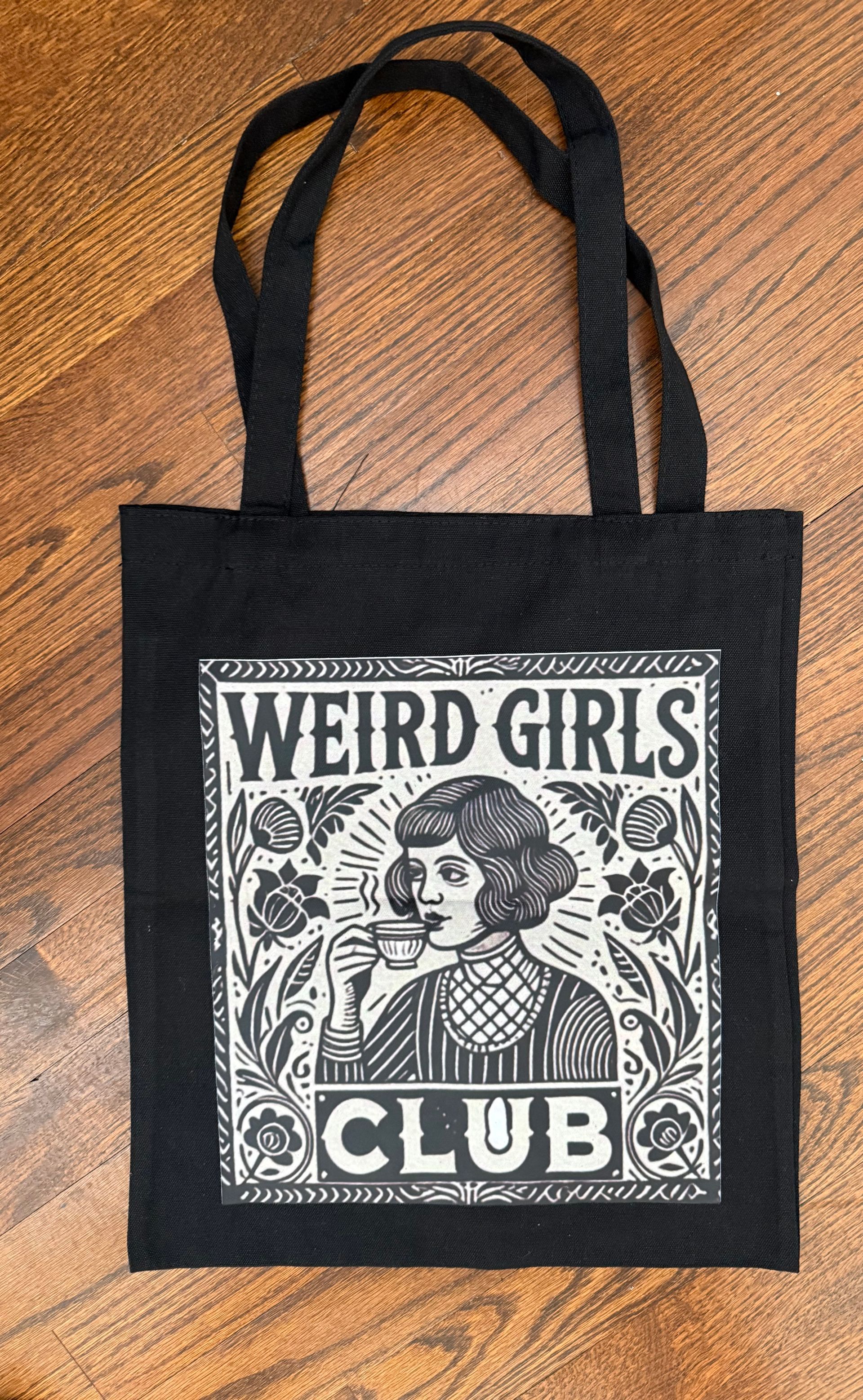 Black tote bag with “WEIRD GIRLS CLUB” graphic on a wooden floor
