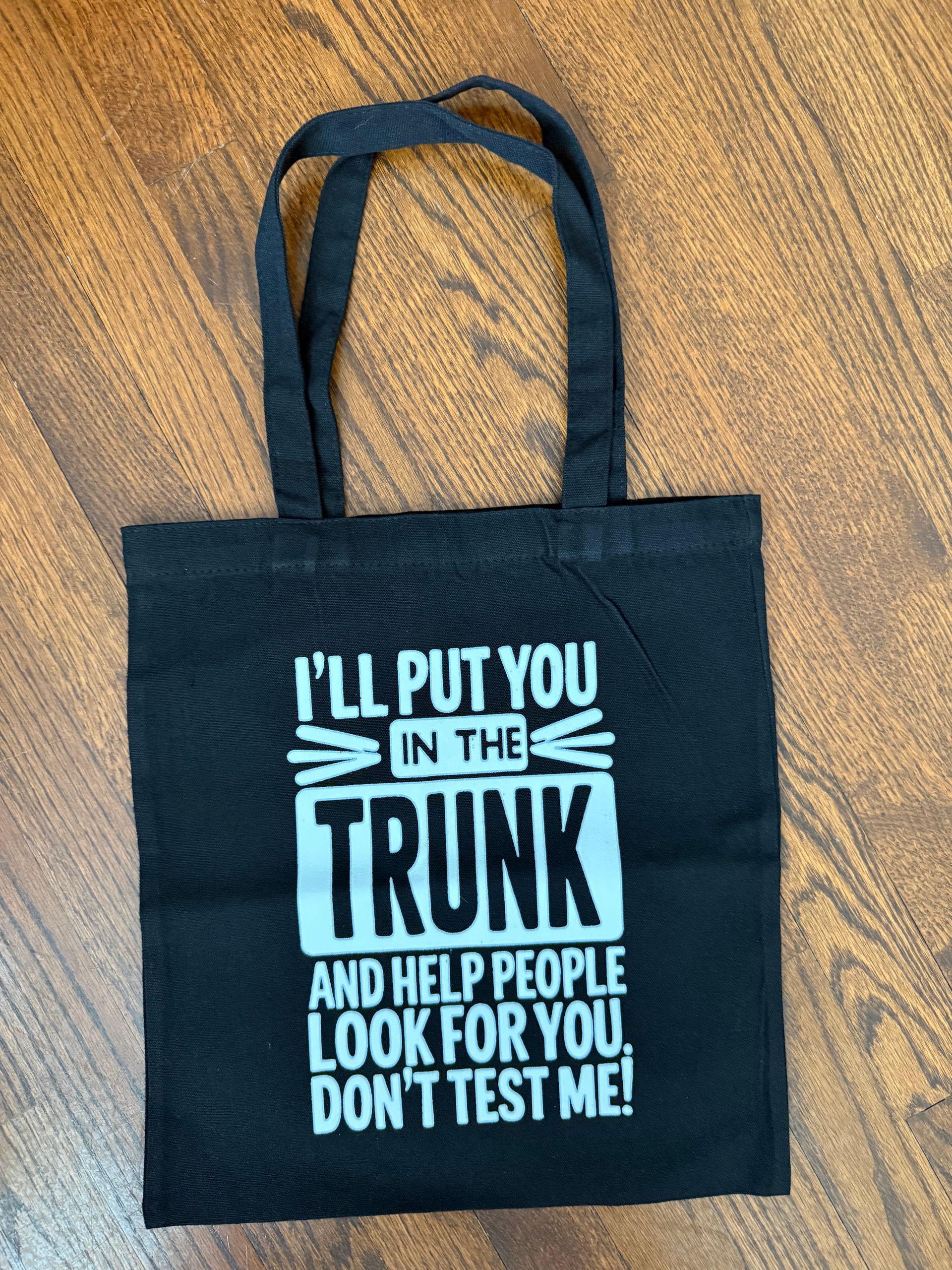 Black tote bag with white text saying, “I’ll put you in the trunk and help people look for you. Don’t test me!”