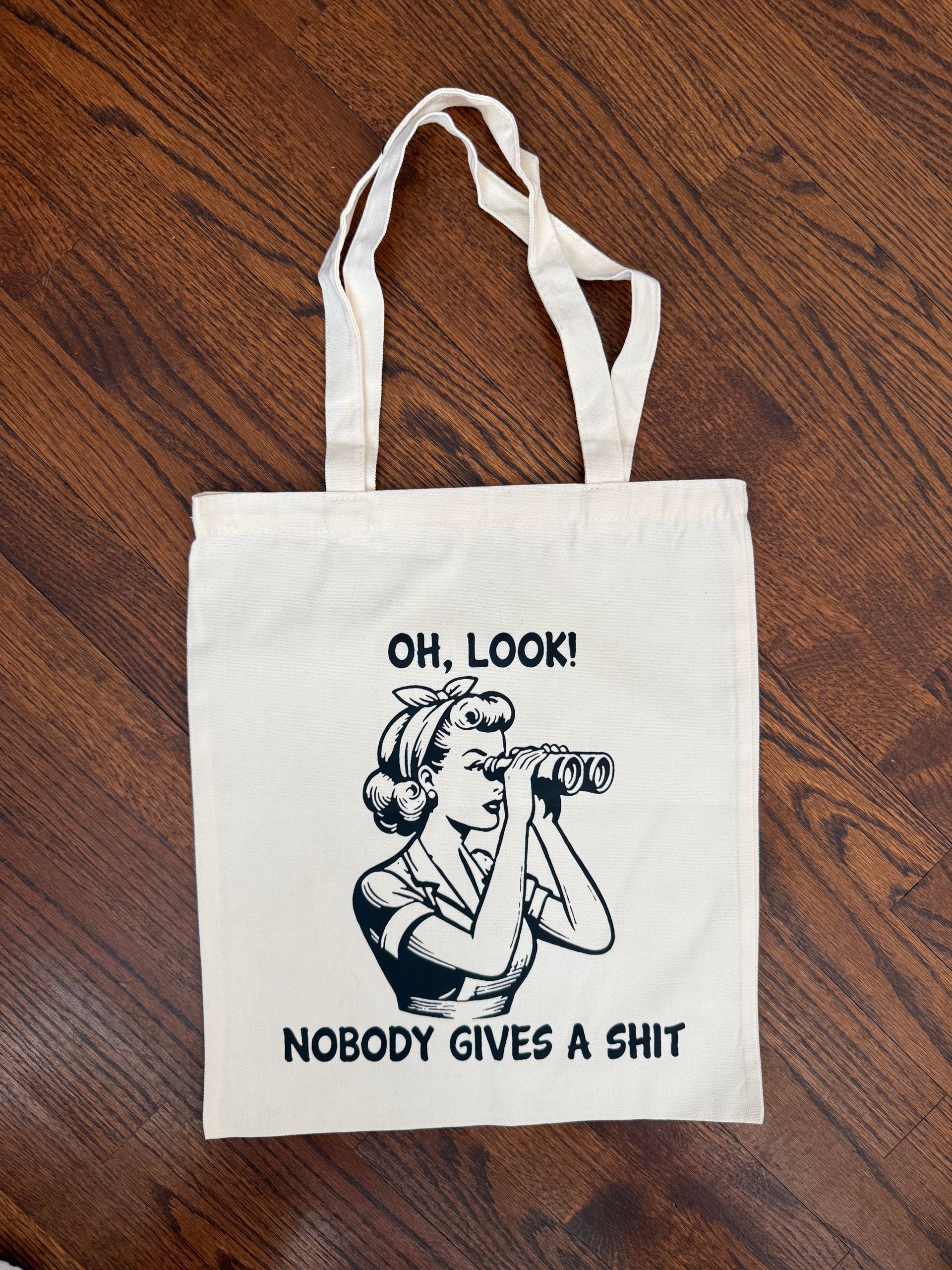 White tote bag on a wooden floor with a cartoon woman and text reading “OH, LOOK! NOBODY GIVES A SHIT.”