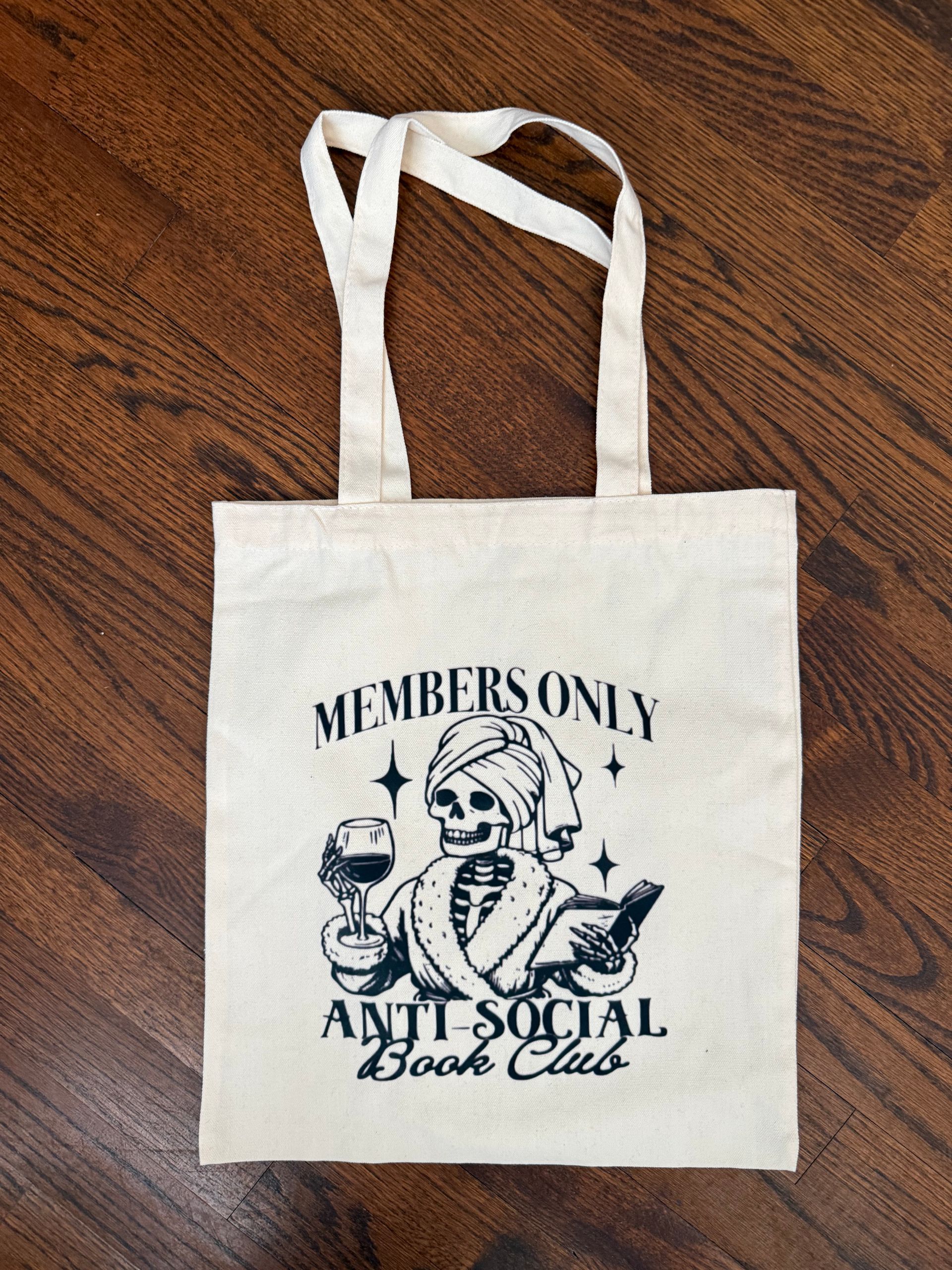 Cream tote bag with “Members Only Anti-Social Book Club” graphic on a wooden floor