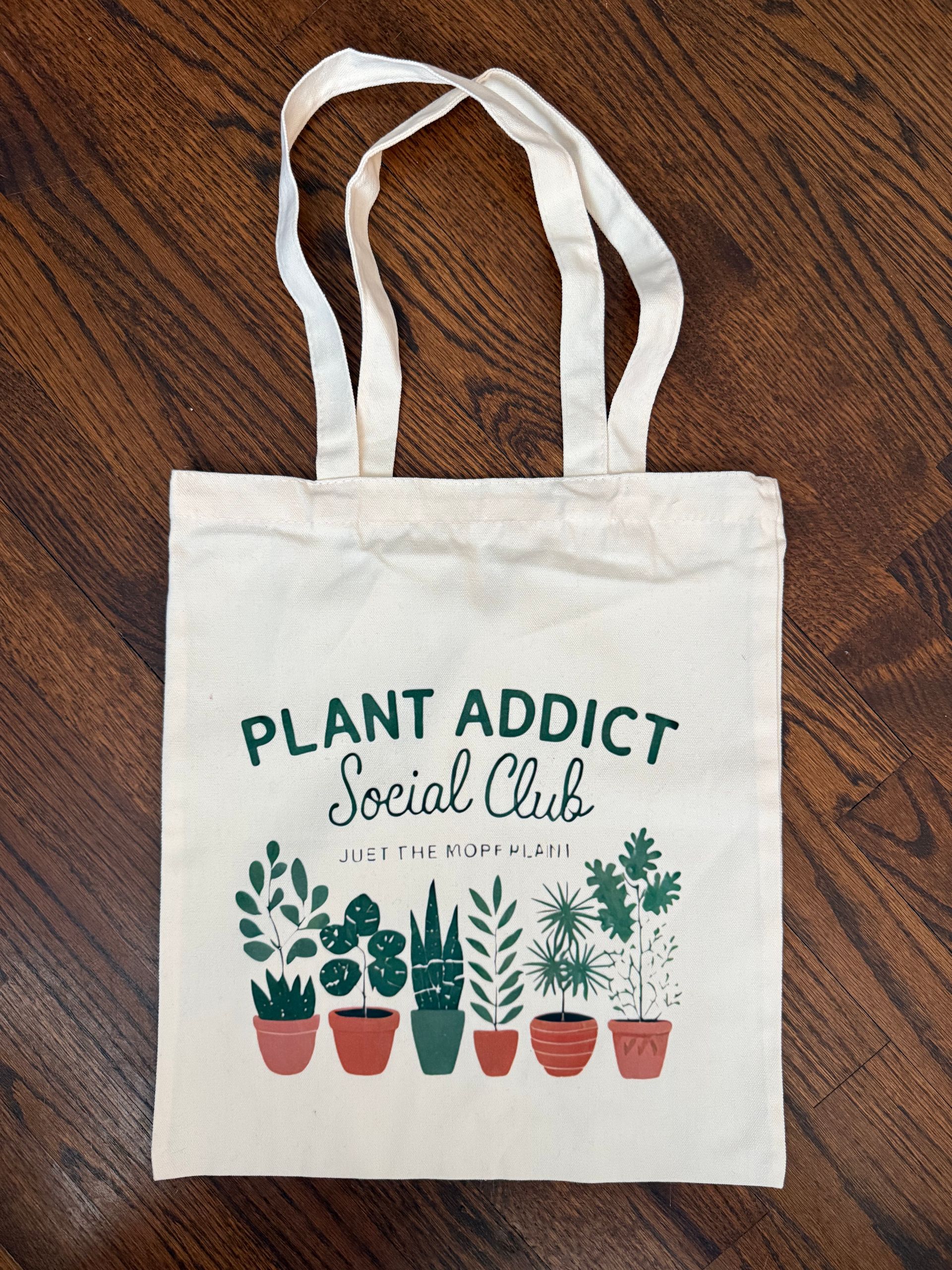 Canvas tote bag reading “Plant Addict Social Club” with potted plant illustrations on a wooden floor