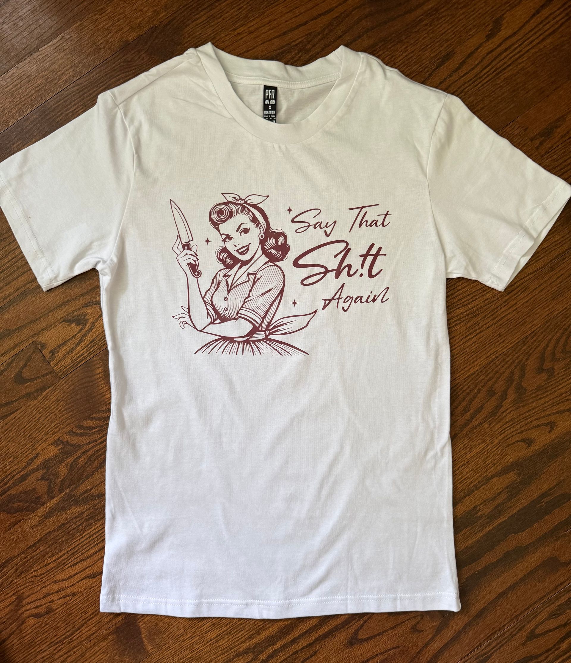 White T-shirt with a dark red graphic and text printed on the front, laid on a wooden floor.
