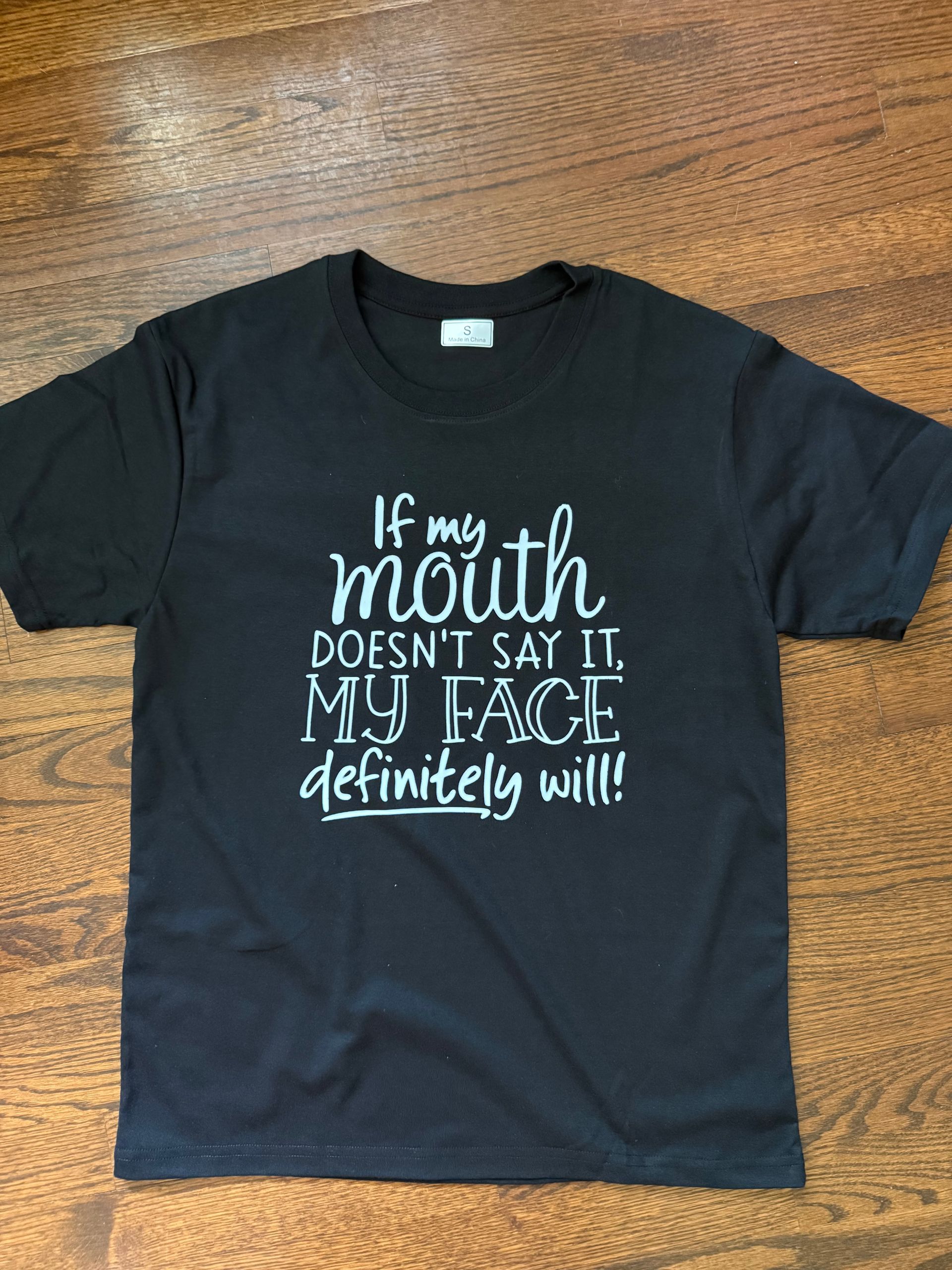 Black T-shirt with white text on a wooden floor: “If my mouth doesn’t say it, my face definitely will.”
