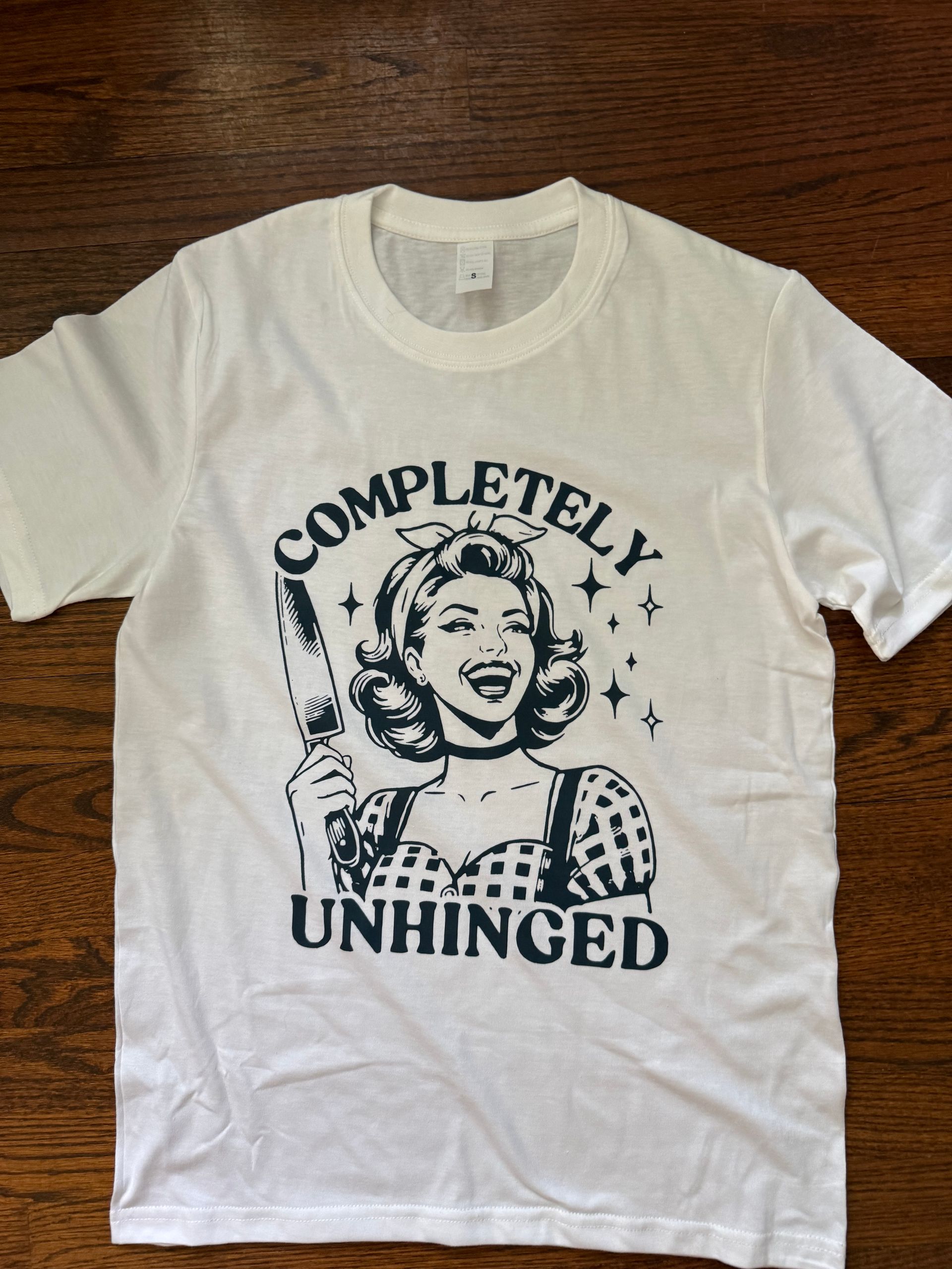 White T-shirt with “COMPLETELY UNHINGED” text and a retro woman graphic on a wooden surface