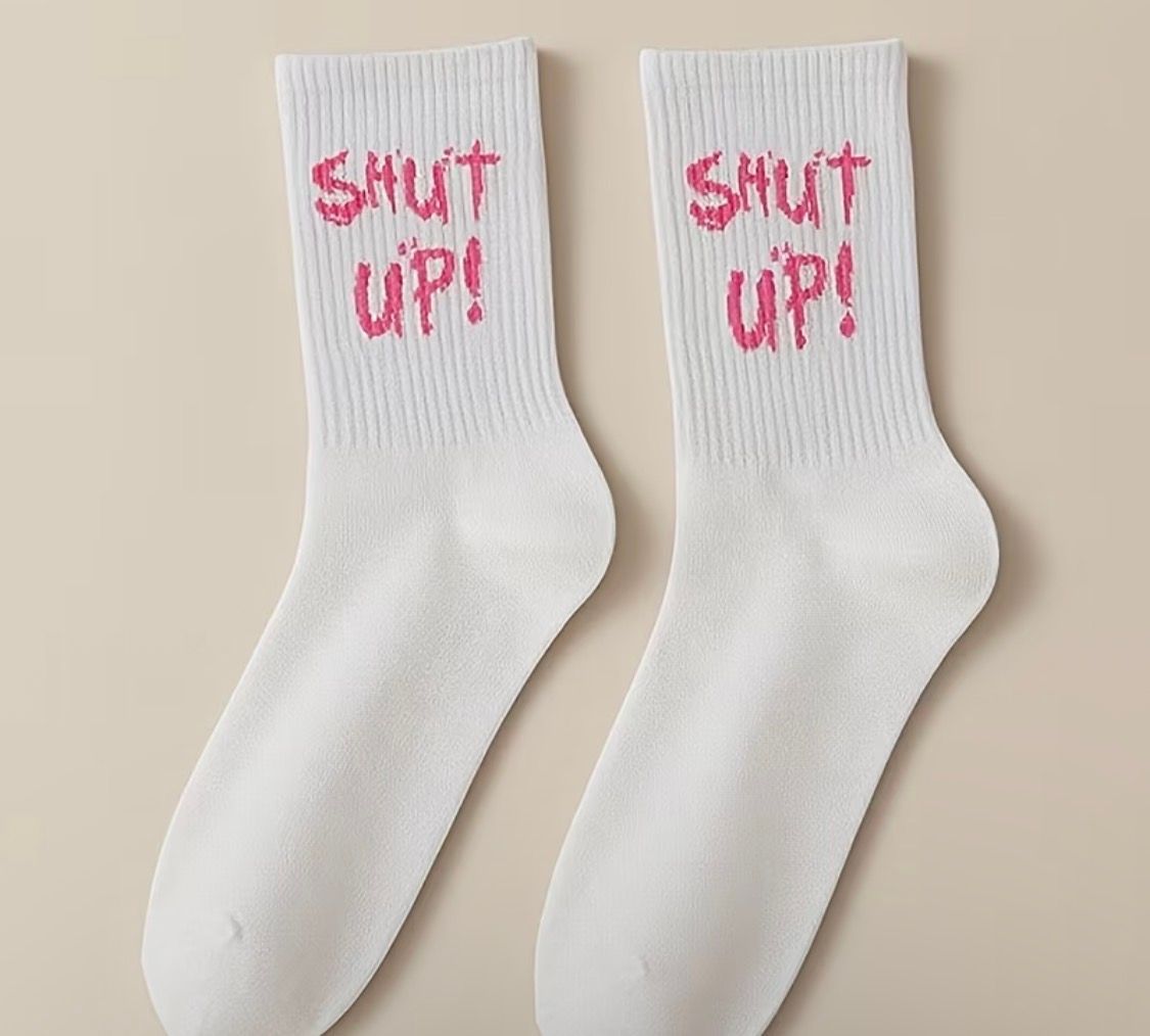 Pair of white socks with pink “shut up!” text on a beige background