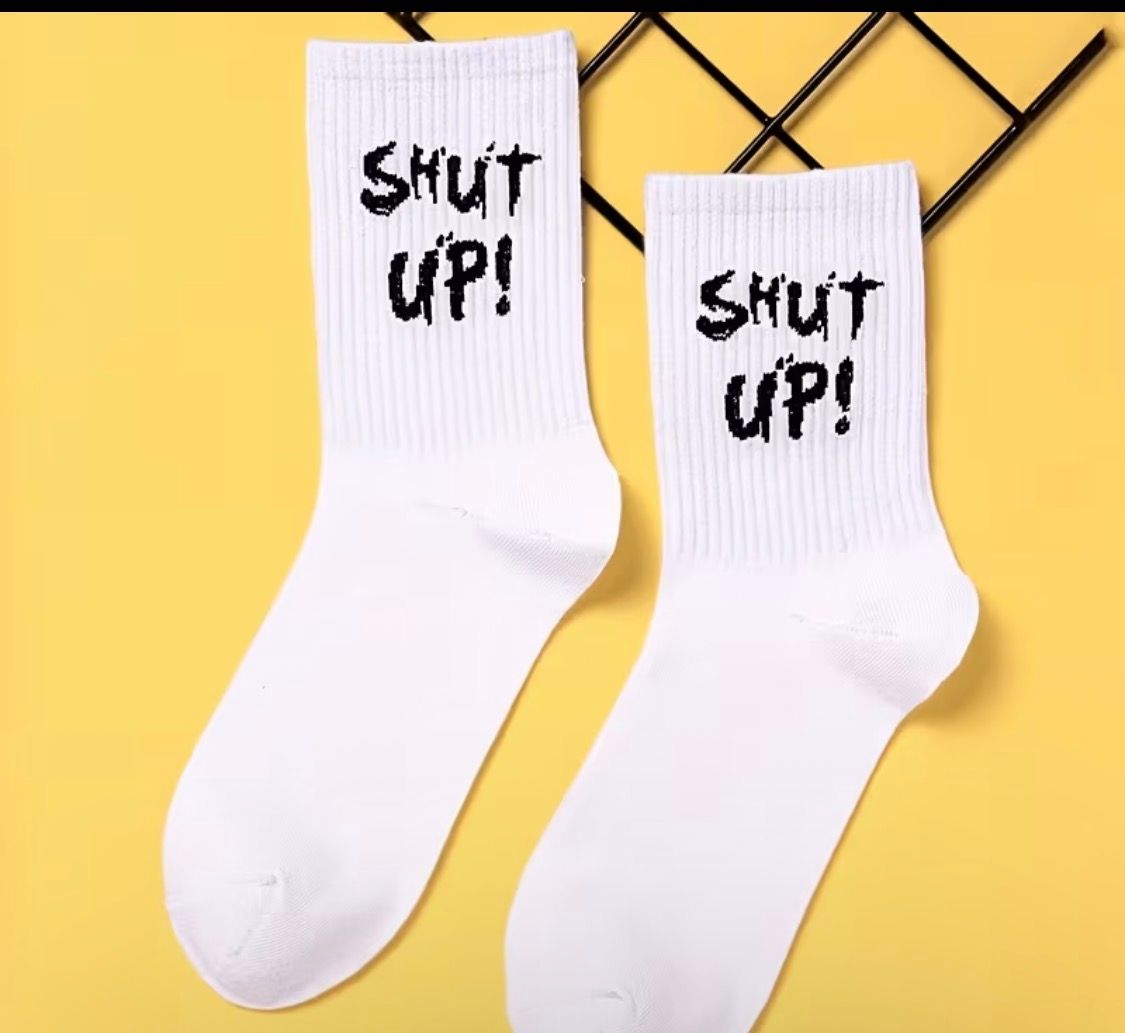 Pair of white socks with pink “shut up!” text on a beige background