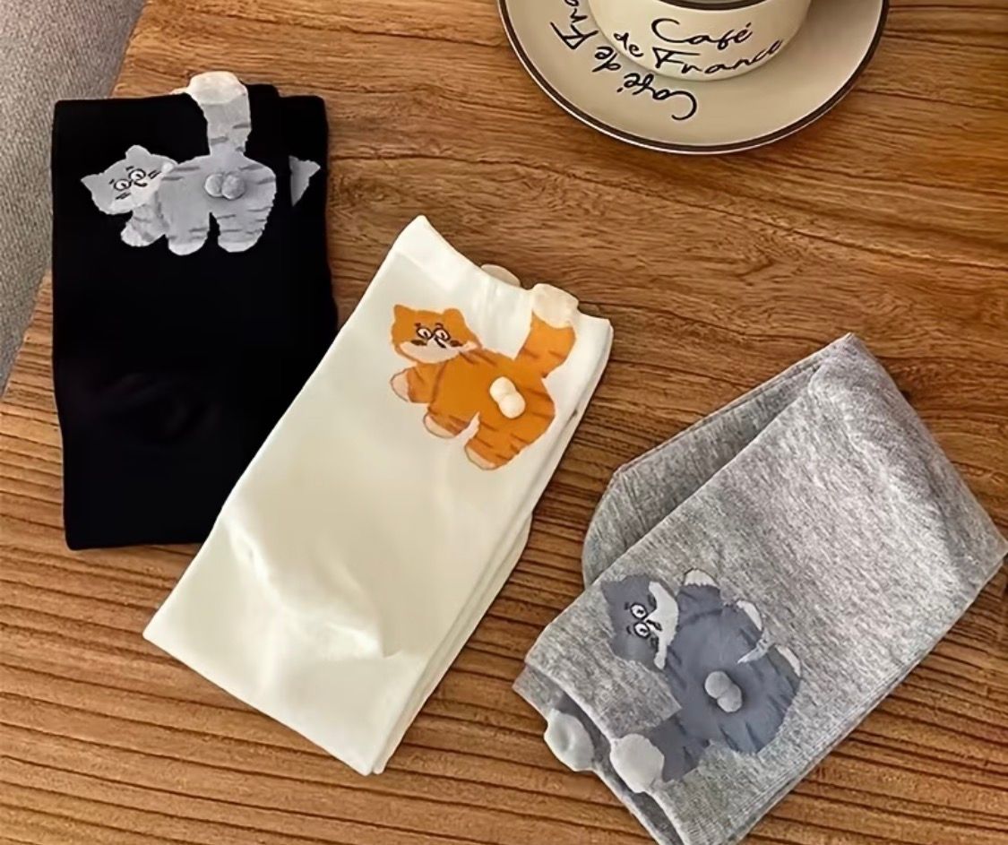 Three folded socks in black, cream, and gray with cat designs on a wooden table.