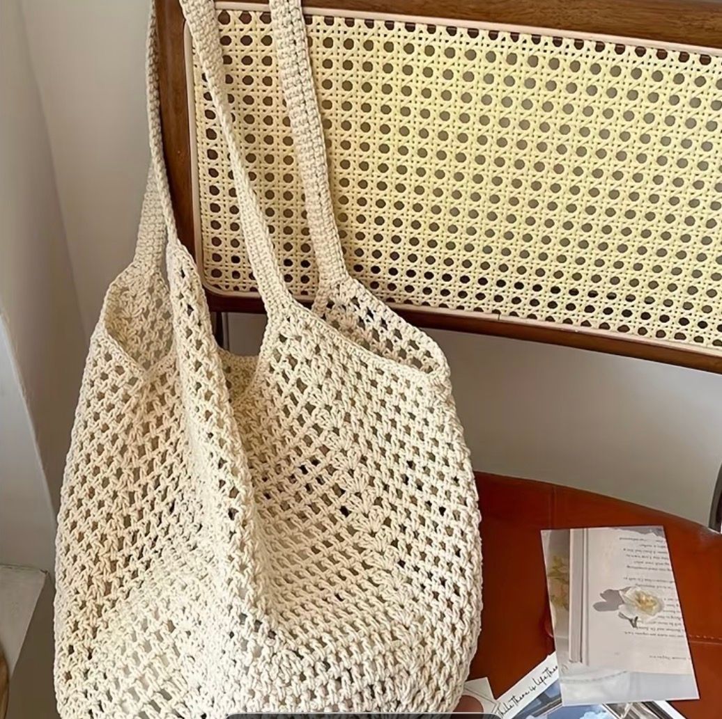 Cream crochet tote bag hanging on a chair beside a small book or notebook on a wooden surface