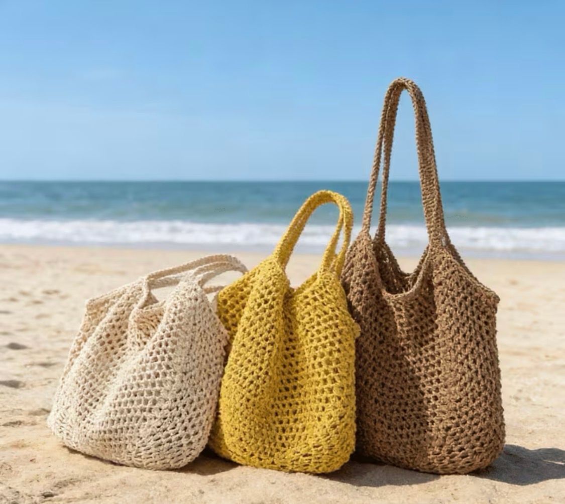 Three woven beach tote bags in cream, yellow, and tan on sandy shore by the ocean
