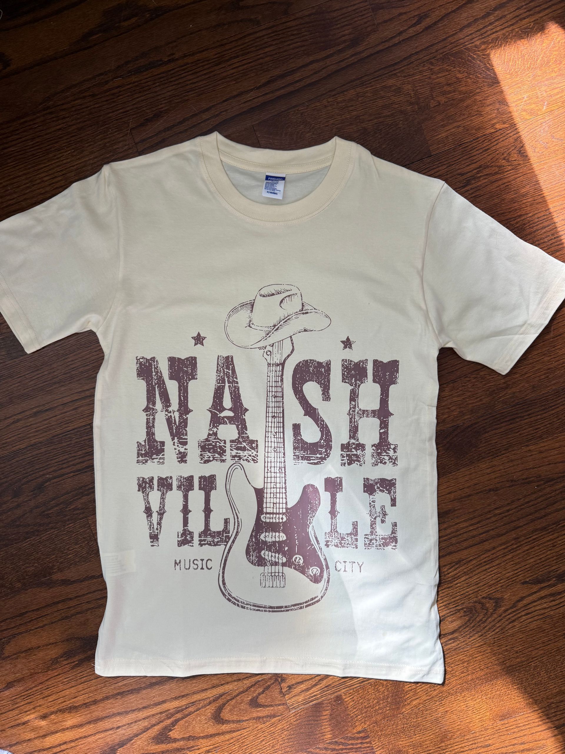 Cream T-shirt with purple “NASHVILLE” graphic laid flat on a wooden floor.