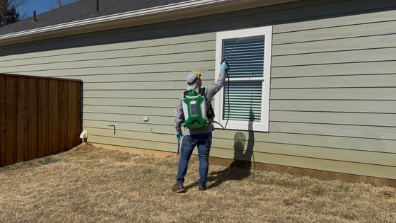 Worker cleaning a house window with a pressure washer beside beige siding and a wooden fence.