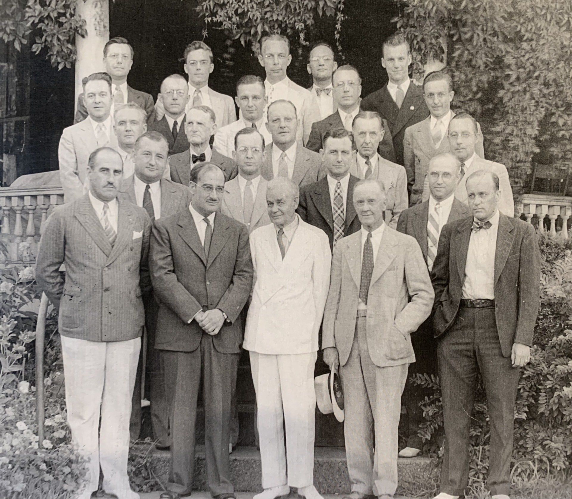 Group of men posing for a photo on steps outside. Mostly wearing suits, some smiling.