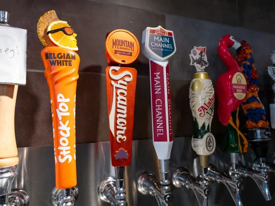 Close-up of beer tap handles; Shock Top Belgian White, Sycamore, Main Channel, Alan's Lake, and another with a pink flamingo.