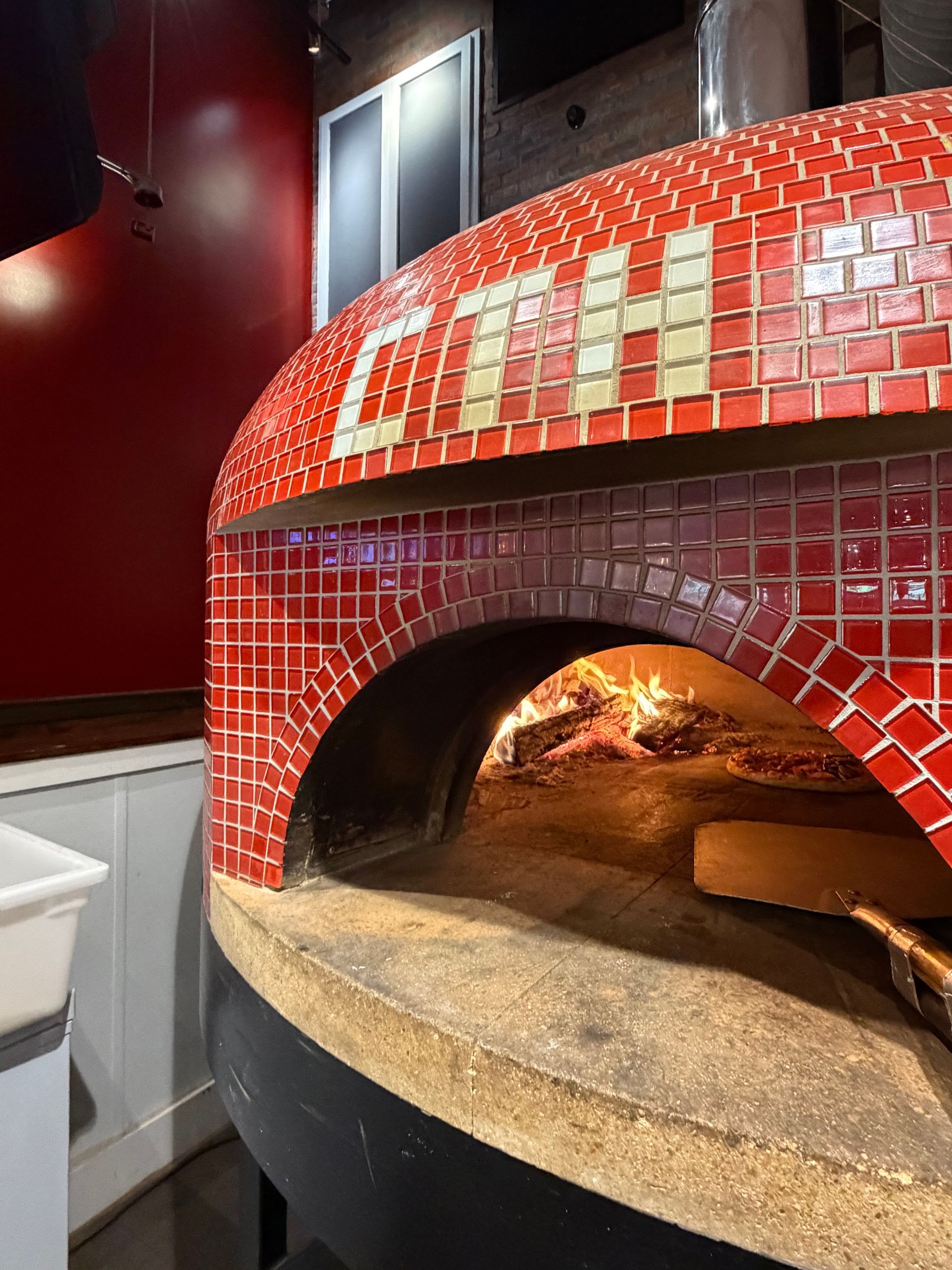 A red tiled wood-fired pizza oven with "CTH" in white tiles above its arched opening, showing flames inside.