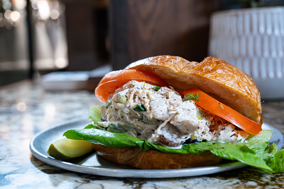 Tuna salad sandwich with tomato and lettuce on a plate, with a pickle and blurry background.