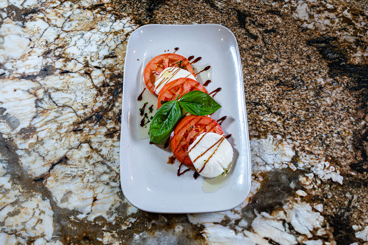 Caprese salad: Tomato and mozzarella slices, fresh basil, drizzled with balsamic glaze and oil on a white rectangular plate.